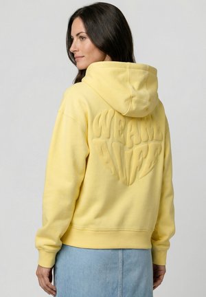 Frau trägt einen gelben Hoodie mit dem erhabenen Schriftzug "WE ARE ALIVE" auf dem Rücken, kombiniert mit einem hellblauen Jeansrock.