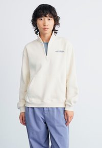 Pull en polaire de couleur crème avec une fermeture éclair quart, une poche kangourou, des poignets côtelés et un petit logo bleu sur la poitrine gauche. Associé à un pantalon bleu clair.