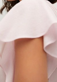 Primo piano del braccio superiore di una persona che indossa un indumento a maniche corte con volant di un rosa chiaro, con capelli scuri parzialmente visibili.