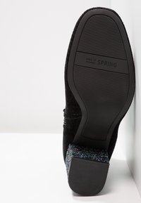 Chaussure en suède noir avec un talon bloc recouvert de paillettes multicolores. La semelle est en caoutchouc texturé avec "CALL IT SPRING" embossé.