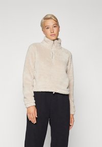 Pull en polaire beige clair, duveteux, avec un col montant, une fermeture éclair courte et des poignets élastiqués. Associé à un pantalon ample sombre.