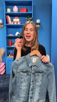 Woman holding a denim jacket on a hanger and a makeup brush, standing in front of blue shelves with decorative items and plants.