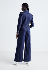 Ținută din denim, compusă dintr-o cămașă cu mânecă lungă și pantaloni evazați. Material de culoare albastru închis cu cusături contrastante și o croială lejeră.