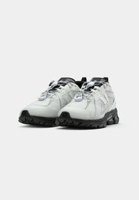 Scarpe sportive bianche e nere con tessuto testurizzato, dettagli in rete e una comoda suola in gomma spessa con un motivo di battistrada. Le stringhe sono elastiche.