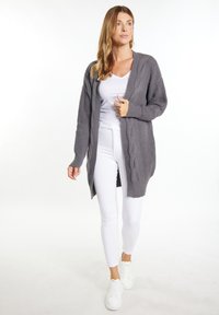 Grauer gestrickter Cardigan mit Zopfmuster, offenem Vorderteil und gerippten Bündchen, kombiniert mit einem weißen Oberteil und Skinny-Jeans. Die Schuhe sind weiße Sneaker.