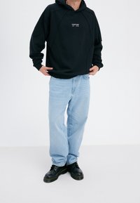 Sudadera negra de gran tamaño con un logo en el frente, combinada con jeans rectos de mezclilla de color azul claro y botas negras con cordones. Tejido suave y corte relajado.