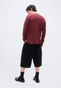 Νέος άνδρας που στέκεται με την πλάτη γυρισμένη, φορώντας μια μακρυμάνικη πουκάμισο σε σκούρο βουργουνδί με υφή, μαύρα φαρδιά σορτς μήκους μέχρι το γόνατο, μαύρες κάλτσες και μαύρα μοκασίνια.