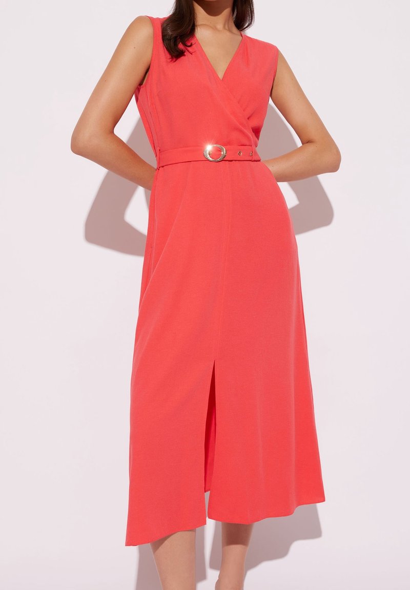 Robe midi corail sans manches avec col en V, taille ceinturée, fente avant, portée par une personne debout les mains sur les hanches sur fond blanc.