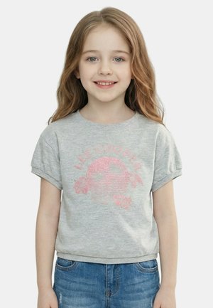 Jeune fille aux longs cheveux châtain clair portant un t-shirt gris Lee Cooper et un jean en denim bleu, souriant à la caméra.