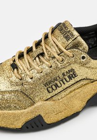 Zlaté třpytivé tenisky s černými akcenty, featuring objemnou podrážkou, texturovaným povrchem a tkaničkami. Výrazné logo "Versace Jeans Couture".