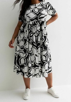 Vrouw in een zwart-witte bloemenmidi-jurk met korte pofmouwen en witte sneakers tegen een effen witte achtergrond.