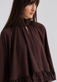 Blouse en soie marron avec un col haut, fermetures à boutons sur le devant et un ourlet froncé. Le tissu semble lisse et léger.