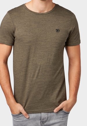Homme portant un t-shirt à manches courtes vert olive uni avec un petit logo noir sur la poitrine gauche, les mains dans les poches d'un jean gris clair.