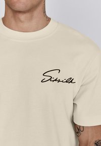Camiseta de algodón color crema con cuello redondo y mangas cortas. Presenta un logotipo de cursiva negra en el lado izquierdo del pecho. Textura suave.