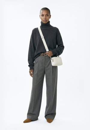 Persoană purtând un pulover golf gri închis, pantaloni gri largi, pantofi maro cu vârf ascuțit, curea cu print leopard și geantă albă de tip crossbody pe fundal alb.