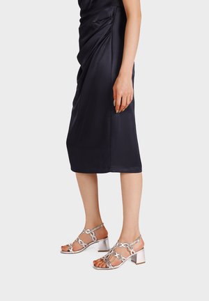 Donna che indossa un abito midi nero e sandali argento con tacco a blocco e punta aperta, con cinturini decorativi simili a catene, in piedi su uno sfondo bianco.