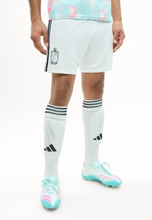 Mann trägt weiße Fußballshorts mit Wappen, weiße kniehohe Socken mit schwarzen Streifen und Schienbeinschoner sowie weiße Fußballschuhe mit türkisfarbenen und pinken Akzenten.