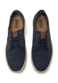 Zapatos de lona azul marino con parte superior perforada, acentos de cuero marrón y suelas con textura similar al arpillera. Presenta cinco ojetes y una plantilla de corcho.