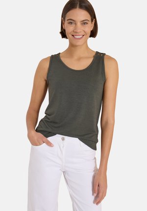 Femme aux cheveux courts bruns souriante, portant un haut sans manches vert foncé et un pantalon blanc, avec une main dans la poche.