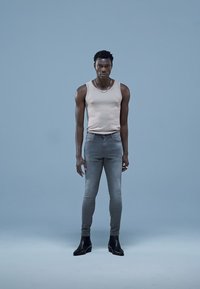 Modell iført en beige singlet og smale grå jeans, kombinert med svarte ankelstøvler. Glatte teksturer mot en lyseblå bakgrunn.