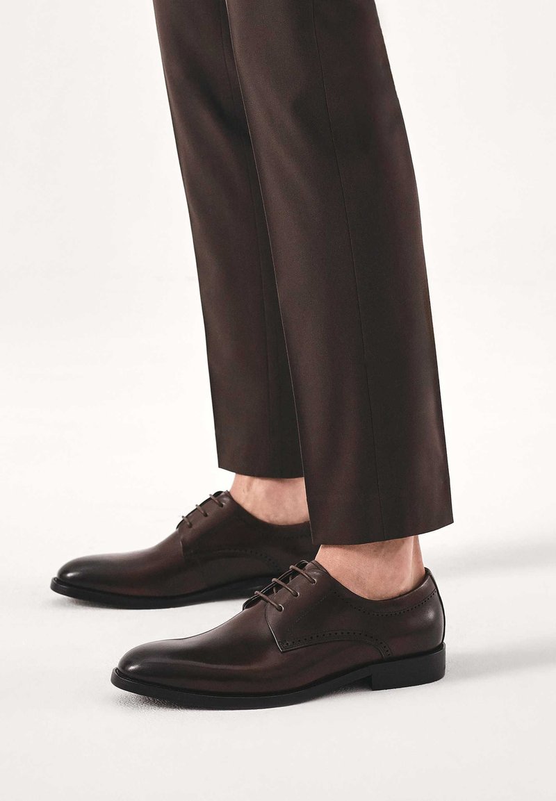 Scarpe eleganti marroni in pelle con finitura liscia, con design con lacci e cuciture decorative lungo i lati, abbinate a pantaloni scuri.