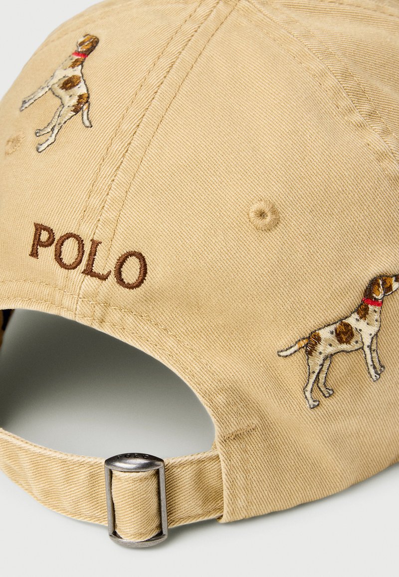 Beige Baseballkappe mit gestickten braunen und weißen Hunden, braunem "POLO"-Schriftzug und verstellbarem Metallverschluss am Hinterkopf.