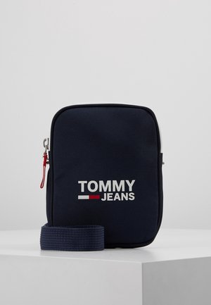 Sac bandoulière compact Tommy Jeans bleu marine avec logo blanc et rouge, tirette de fermeture éclair rouge, et sangle bleu marine assortie sur surface blanche.