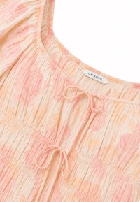 Blouse texturée à motifs floraux rose clair et pêche avec col élastique, deux liens à nouer à l'avant et étiquette portant la mention « OH APRIL » à l'intérieur du col.