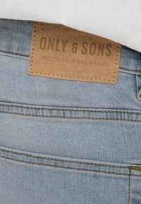 Jeans de mezclilla azul claro con una superficie texturizada, costuras amarillas y una etiqueta de cuero marrón con "ONLY & SONS" y "PASIONADOS CREADORES DE JEANS".