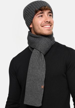 Ensemble bonnet et écharpe en tricot côtelé gris, fabriqué en tissu doux, avec une accent en cuir sur l'écharpe, porté sur une chemise noire.
