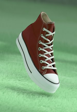 Rode hoge sneakers van canvas met een witte rubberen zool, zwarte accentlijn en witte veters, weergegeven op een groene gestructureerde ondergrond.