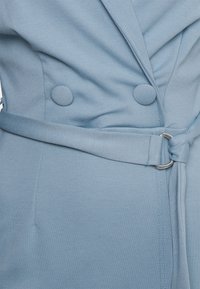 Vaaleansininen takki, joka on valmistettu tekstuuripinnasta ja jossa on tupla-napit, vyötäröllä vyö hopeisella soljella sekä läppäkollarin yksityiskohtia.