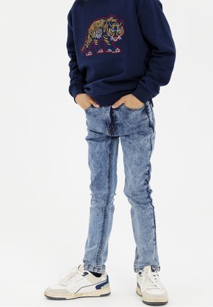 Enfant portant un sweat-shirt bleu marine avec une broderie de tigre, un jean bleu clair, des baskets blanches, debout les mains dans les poches sur un fond blanc.