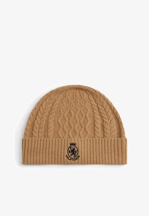 Tan gebreide beanie met een kabelpatroon en een geribde rand. Inclusief een zwart geborduurd logo aan de voorkant. Zachte textuur.