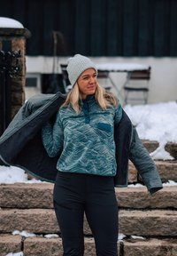 Pull en polaire à motifs bleus avec une fermeture éclair quart et une poche poitrine, superposé sous une veste grise, porté avec un pantalon noir, sur des marches couvertes de neige.