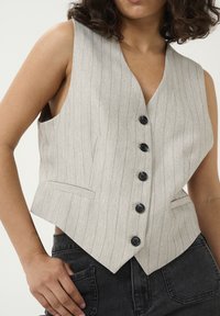 Gilet rayé gris clair avec un décolleté en V profond, six boutons noirs et des poches latérales, associé à un jean en denim foncé. Tissu texturé.