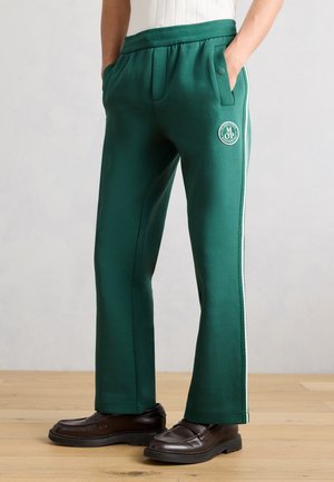 Osoba ubrana w zielone spodnie dresowe z bocznym paskiem i logo, połączone z brązowymi skórzanymi lordami, stojąca na drewnianej podłodze.