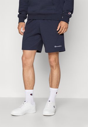 Homme portant un short Champion bleu marine, un sweat assorti, des chaussettes blanches avec le logo Champion et des baskets blanches, debout devant un fond uni.