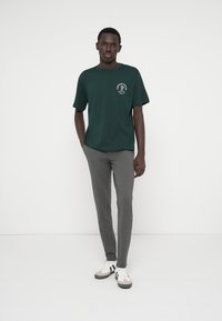 Tume roheline puuvillane T-särk lühikeste varrukatega, ümar kaelus ja valge tekstiga logo. Kandev hallide kitsaste pükste ja mustade-valgete tossudega.