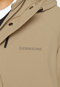 Beżowa kurtka o gładkiej fakturze, z kapturem, zapinana na zamek, z napisem "DIDRIKSONS" w kolorze szarym. Ostry, nowoczesny design.
