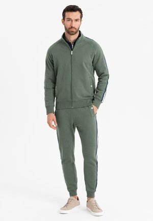 Mand står iført olivengrøn lynlåstrøje og matchende joggingbukser med sidestriber, beige sneakers, hænderne i lommerne.