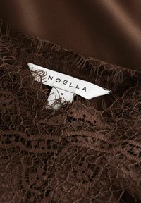 Bruine kanten top met ingewikkelde patronen, gelaagd over satijnen stof. Heeft een label met de tekst "NOELLA," maat "S," en details in witte letters.