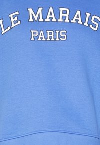 Sudadera azul claro con texto blanco en negrita que dice "LE MARAIS" y "PARÍS" en el pecho. Material de algodón suave, corte estándar.