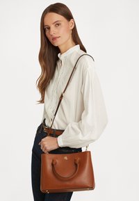 Brown leather handbag with rounded handles, gold hardware accents, and a detachable shoulder strap, complemented by a subtle embossed logo.