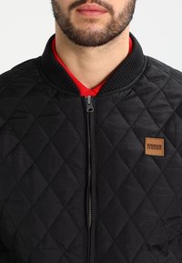 Bearded man wearing a black quilted zip-up jacket with ribbed collar and a red shirt underneath. Urban Classics logo patch on chest.