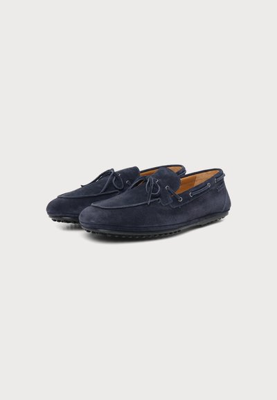 Paire de mocassins en daim bleu marine avec détails de lacets et semelles cloutées en caoutchouc, présentée sous un léger angle sur un fond uni.