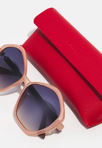 Ochelari de soare hexagonali cu ramă de culoare roz deschis și lentile întunecate cu gradient, împreună cu o husă texturată din piele roșie împăturită cu "GUESS."