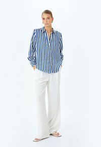 Camicetta a righe blu e bianche con maniche lunghe, abbinata a pantaloni bianchi larghi e sandali bassi.