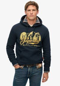 Mørkeblå hættetrøje med en front kangaroo lomme, gyldent grafisk design af bjerge og træer, lagdelt over en plaidskjorte, båret med blå jeans.