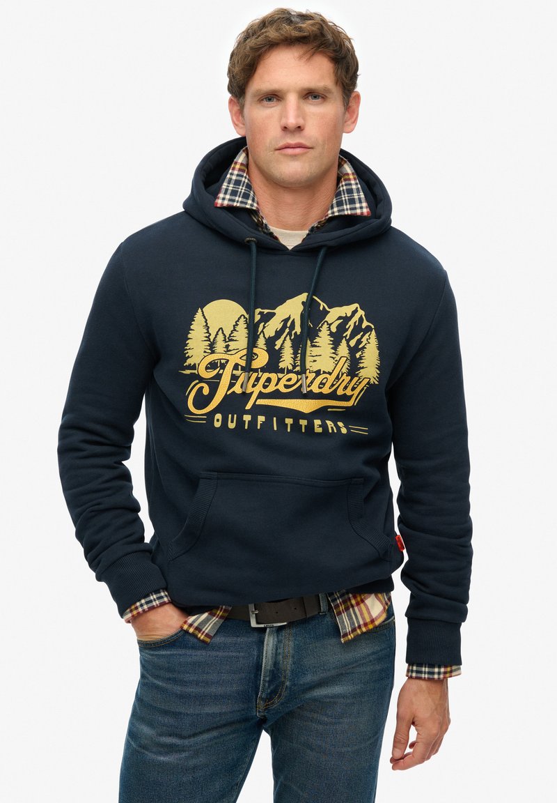 Mørkeblå hættetrøje med en front kangaroo lomme, gyldent grafisk design af bjerge og træer, lagdelt over en plaidskjorte, båret med blå jeans.